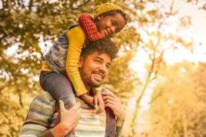 A man carries a child on his shoulders, both smiling, amidst a sunlit park with trees and a warm, golden atmosphere.