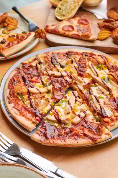 Pizza topped with chicken and drizzled with barbecue sauce, resting on a wooden table alongside additional pizza slices, garlic bread, and cutlery, suggesting a casual dining setup.