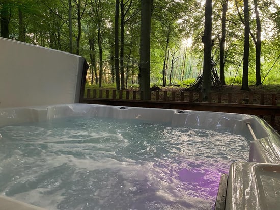 A hot tub bubbles gently in a forest setting, surrounded by lush green trees and dappled sunlight filtering through the canopy.