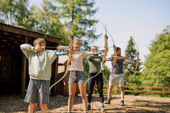 Four people, including two children and two adults, are drawing bows in an outdoor archery range. They are lined up next to a wooden shelter, surrounded by trees under a clear sky.